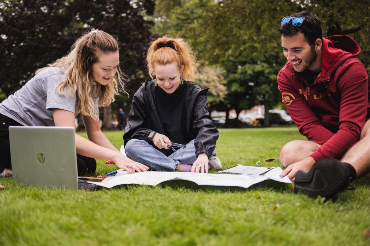 Three people sitting on grass looking at large piece of paper. There is also a laptop in the photo.