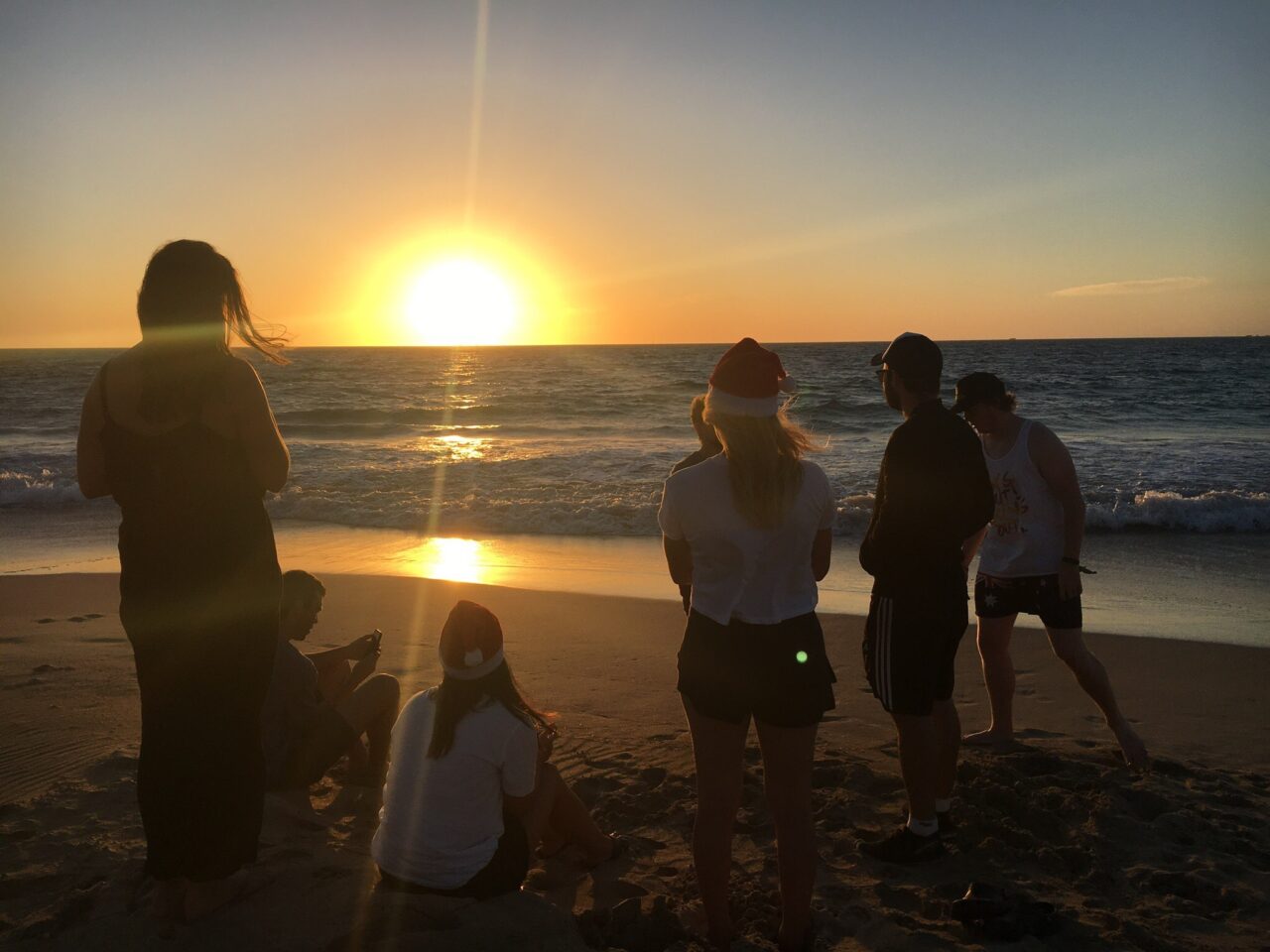 A group of students looking out to a sunset on a beach, two students are wearing Santa hats.