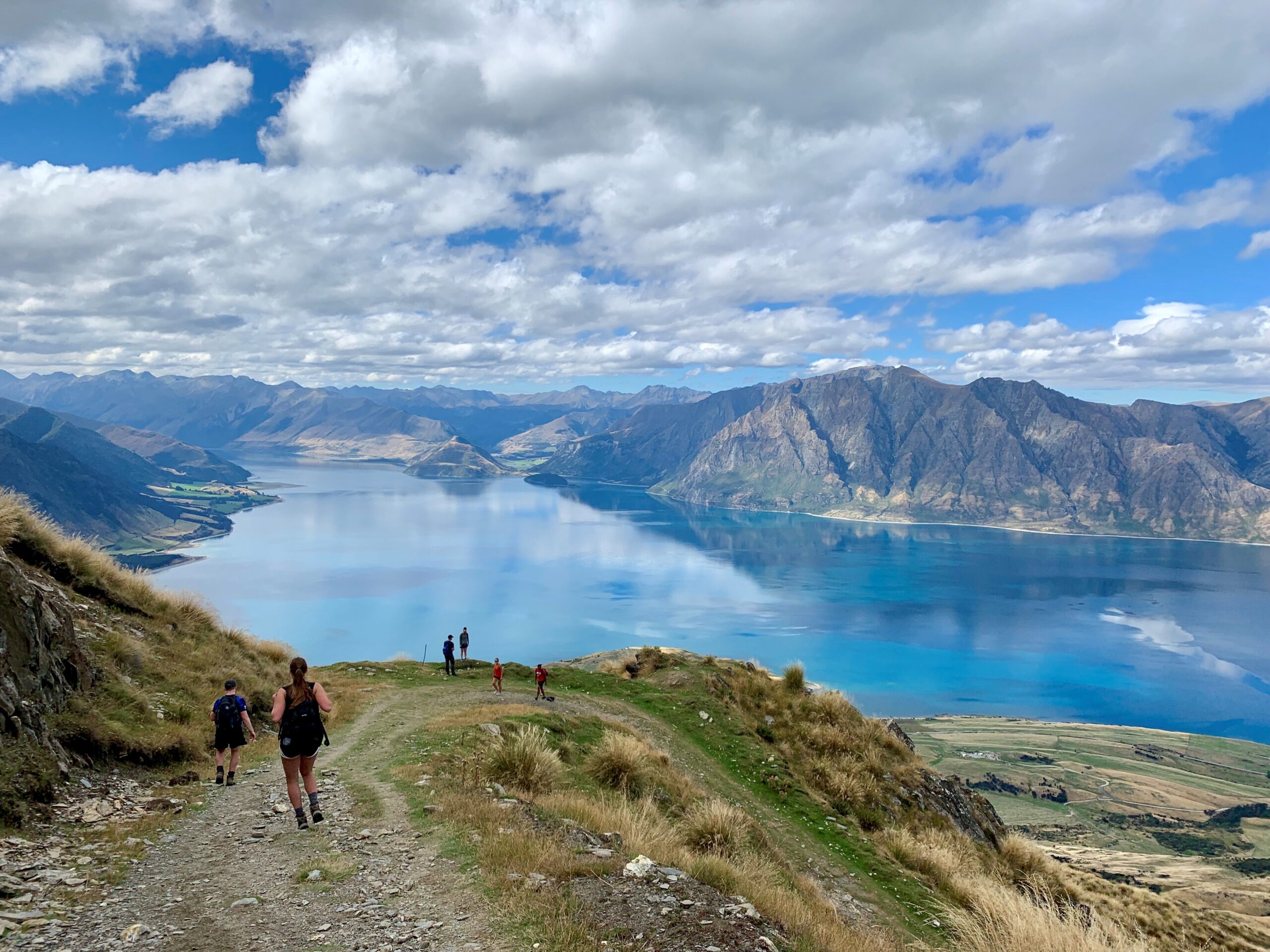 A view of a lake with mountains in the background. There are some people walking down a path and others can be seen in the distance.