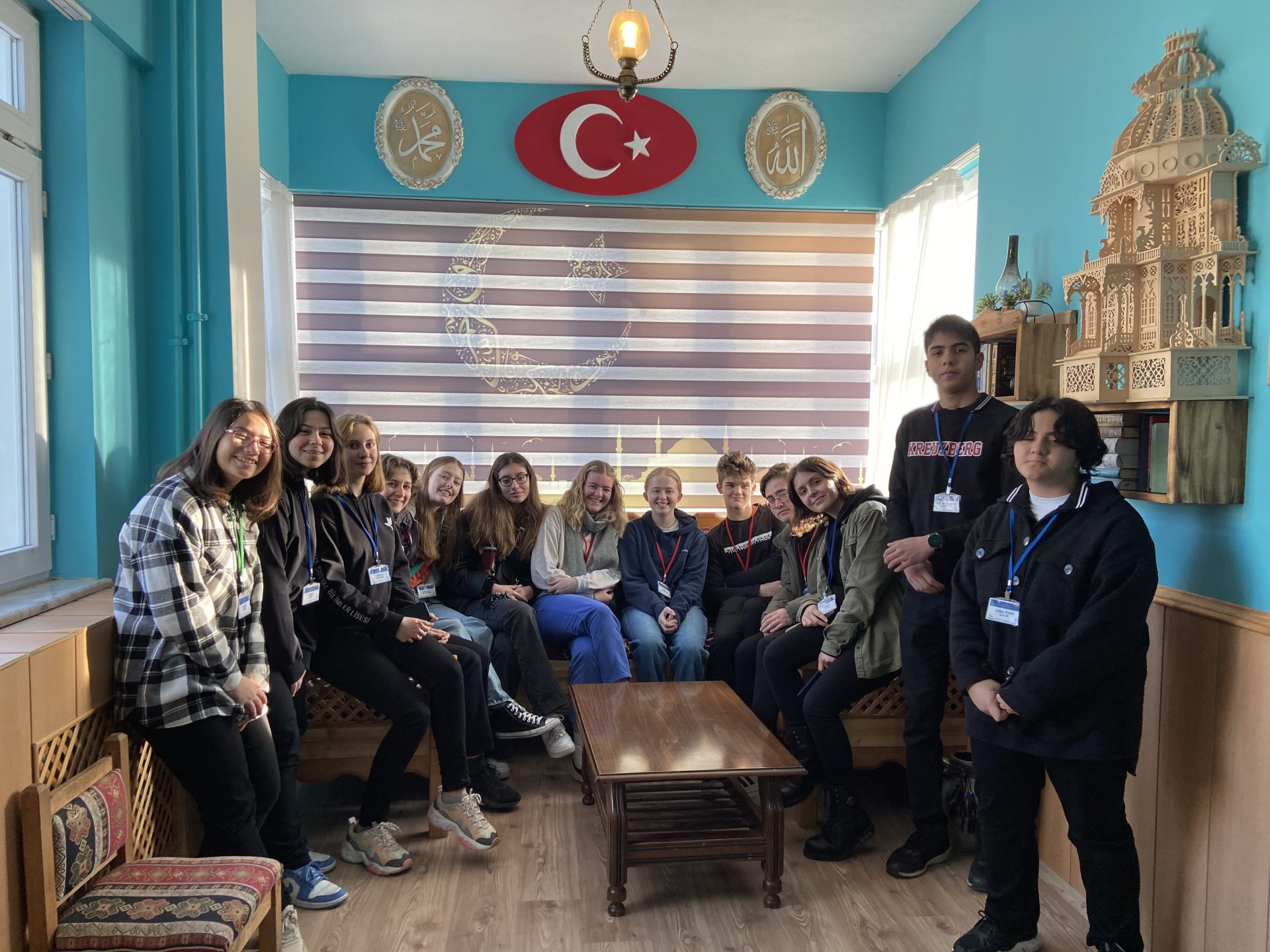 A group of young people (mostly sitting but 2 standing) posing for a photo in a room with blue walls a symbol of Turkey on the wall above them.
