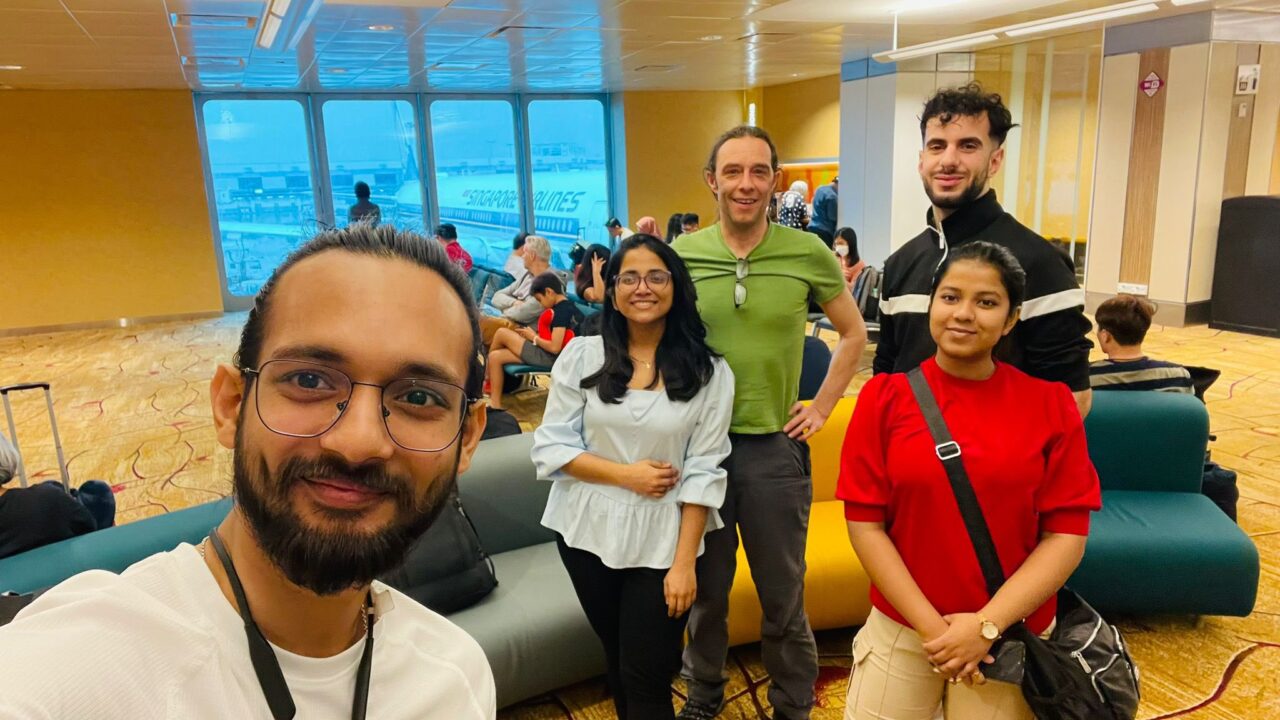Five students smiling at the camera in an airport waiting room with an airplane visible through a window in the background.