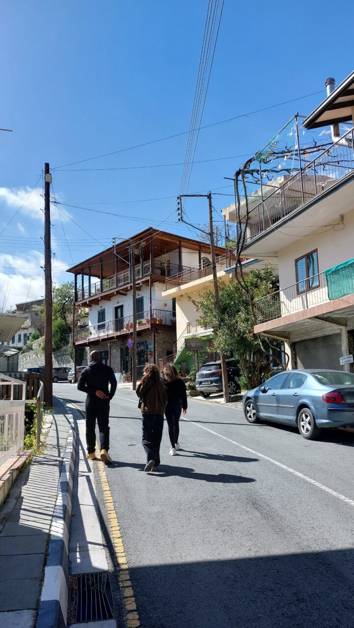 Three people walking up a concrete road on a sunny day. On the right side of the road there are buildings with balconies.