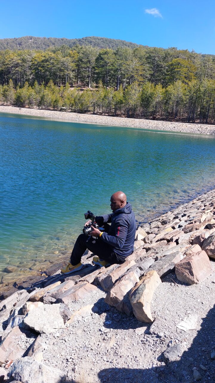 A person sat on a rocky ledge holding a camera by a clear lake, with a backdrop of hills and trees.
