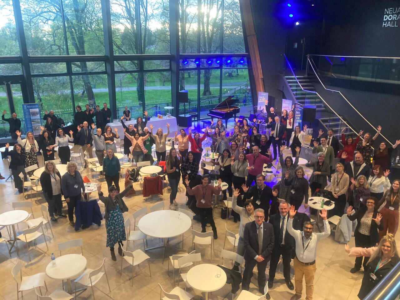 Birds eye view of a conference with a crowd of people looking up and waving. There are round tables with chairs and banners around the outside. A staircase and window looking out to a wooded area are in the background.
