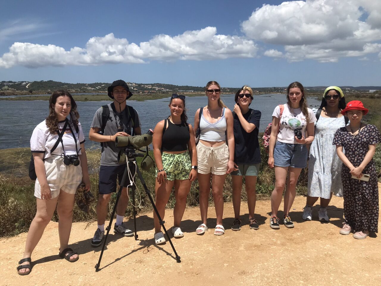 A group of people (with a camera on a tripod) standing in a line. There is water in the background and a blue sky with some cloud.