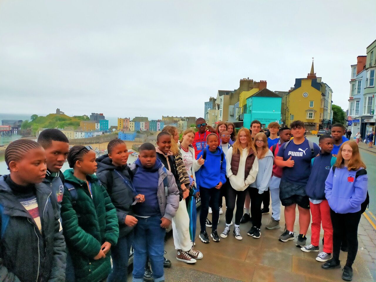 A group of teenagers standing in front of a row of colourful houses in a seaside town.