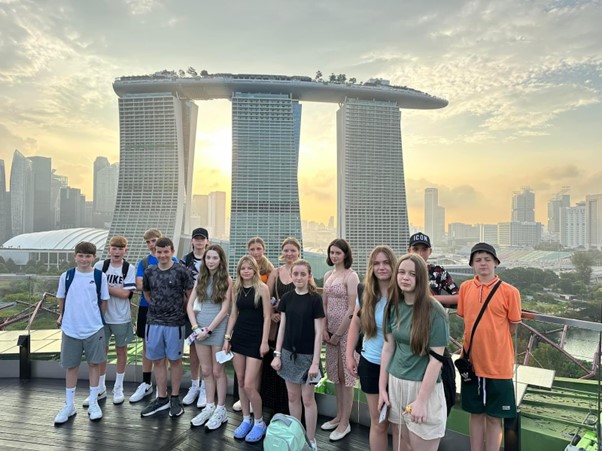 A group of teenagers stand on a platform with an impressive view behind them of three skyscraper buildings with a connecting roof. In the background is a city landscape of further skyscrapers and a sunset sky.