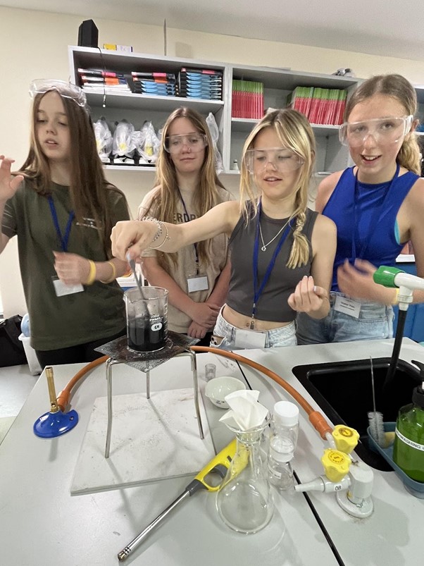 Four teenage girls all wearing protective safety glasses stand together in a classroom setting focusing on a chemistry experiment. One of the girls is placing a spoon into a pot containing dark liquid which sits atop a bunsen burner.