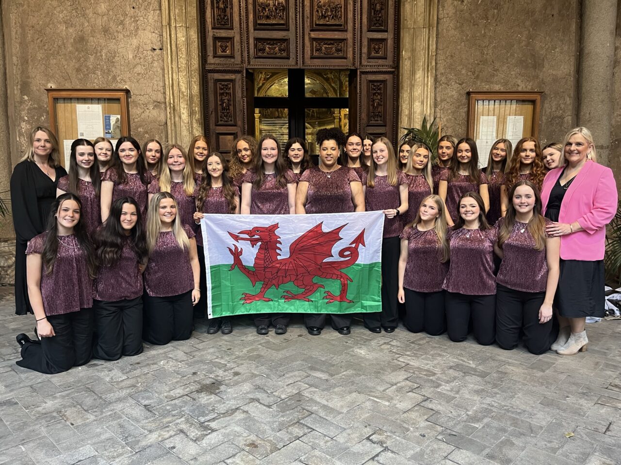 A large group posing for a photo and holding a Welsh flag. They are all wearing similar outfits and appear to be in some sort of gallery/museum.
