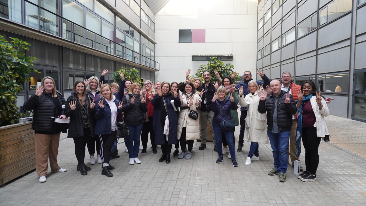 A large group of people standing between two buildings. Most are holding up six fingers.