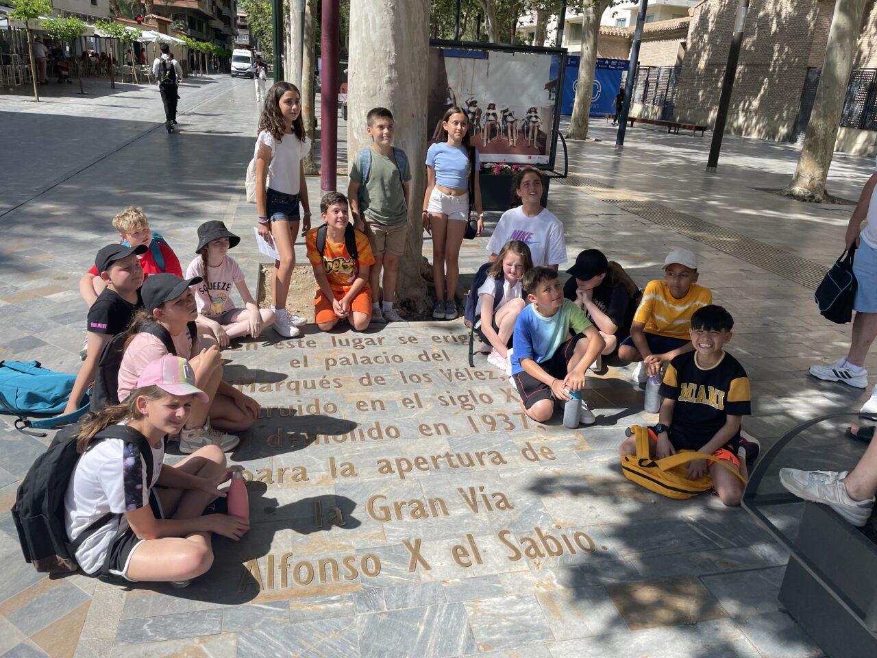 A group of boys and girls sit and stand on the pavement around some paving slabs with golden writing in Spanish. The street can be seen around them which is clean with trees and the sun is shining.