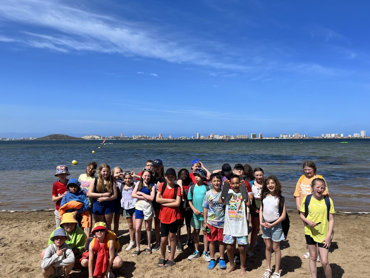 A group of primary school children at the beach on a sunny day. They are standing and kneeling in the sand with the sea visible behind them which leads to a distant landscape of buildings in the background. The sky is blue.