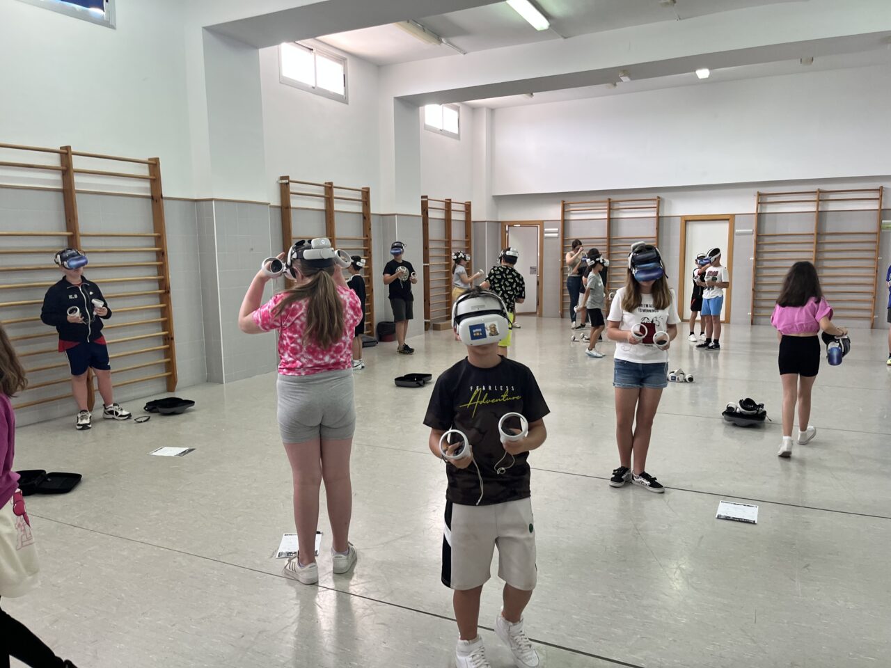 A boy stands in the foreground of a sports hall facing the camera wearing a VR headset. Several boys and girls also wearing VR headsets stand at a distance from each other and facing in different directions in the sports hall.