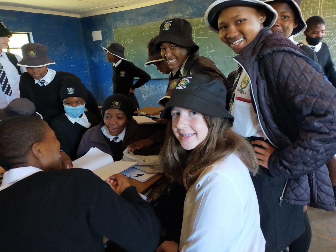 A group of young people in a classroom, where some are smiling at the camera and some are wearing bucket hats. Once person in the background is wearing a mask.