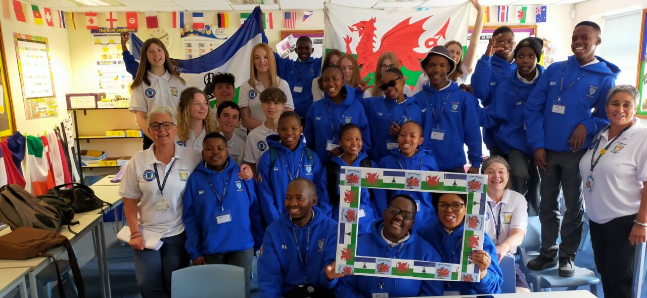 A large group of young people and adults posing for a photo in a classroom setting. Those standing in the back are holding a Welsh and a Lesotho flag.