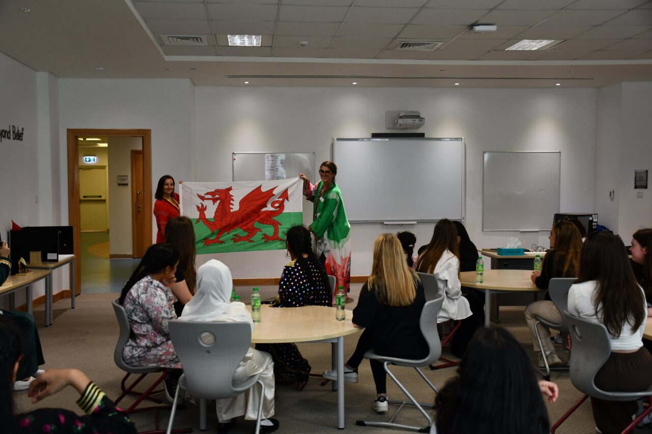 Two female teachers holding the Welsh flag and standing in the front of a classroom of high school girls.