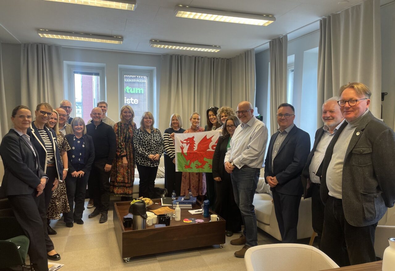 Grŵp mawr o oedolion o'r Ffindir a Chymru yn sefyll o gwmpas bwrdd coffi ac yn gwenu at y camera. Y mae rhai o'r menywod yn gefn y grŵp yn dal baner Cymru.