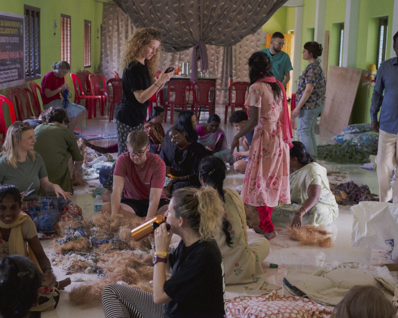 Y tu mewn i ystafell fawr gyda chadeiriau coch o amgylch y tu allan i'r ystafell. Mae llawer o oedolion yn eistedd ar y llawr yn gwneud rhywbeth gyda defnydd tebyg i wlân. Mae eraill yn sefyll i fyny yn sgwrsio. / Inside a large room with red chairs around the outside of the room. Lots of adults are sat on the floor making something with a wool like material. Others are stood up chatting.