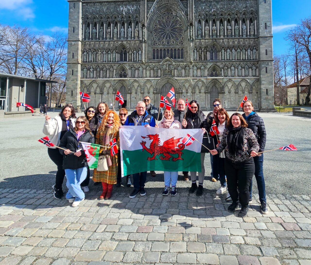 Grŵp o oedolion yn sefyll am lun o flaen eglwys gadeiriol yn Norwy. Mae'r grŵp yn dal baner fawr Cymru a llawer o fflagiau bach Norwyaidd / A group of adults posing for a photo in front of a cathedral in Norway. The group are holding a large Welsh flag and lots of small Norwegian flags