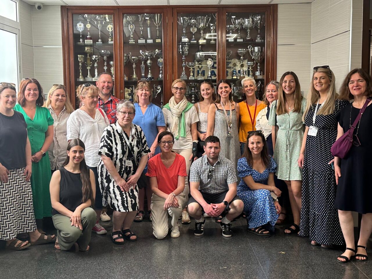 Grŵp mawr o oedolion yn sefyll am lun o flaen cabinet tlws. Mae'r rhan fwyaf yn sefyll ond mae rhai yn penlinio ar gyfer y llun / A large group of adults posing for a photo in front of a trophy cabinet. Most are standing but a few are kneeling for the photo