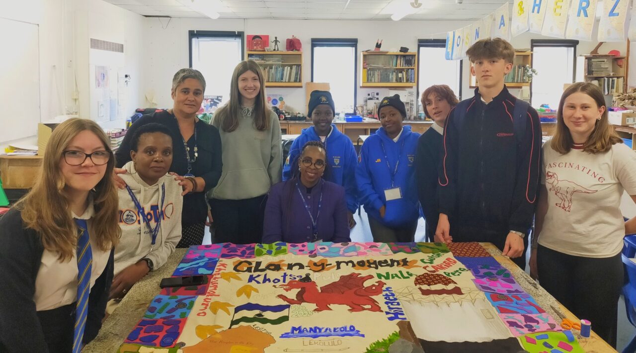 A group of pupils and teachers from Welsh and Lesotho schools gathered around a fabric mural of images that show the schools' shared partnership. There is a Welsh dragon in the centre of the mural and the Lesotho flag.