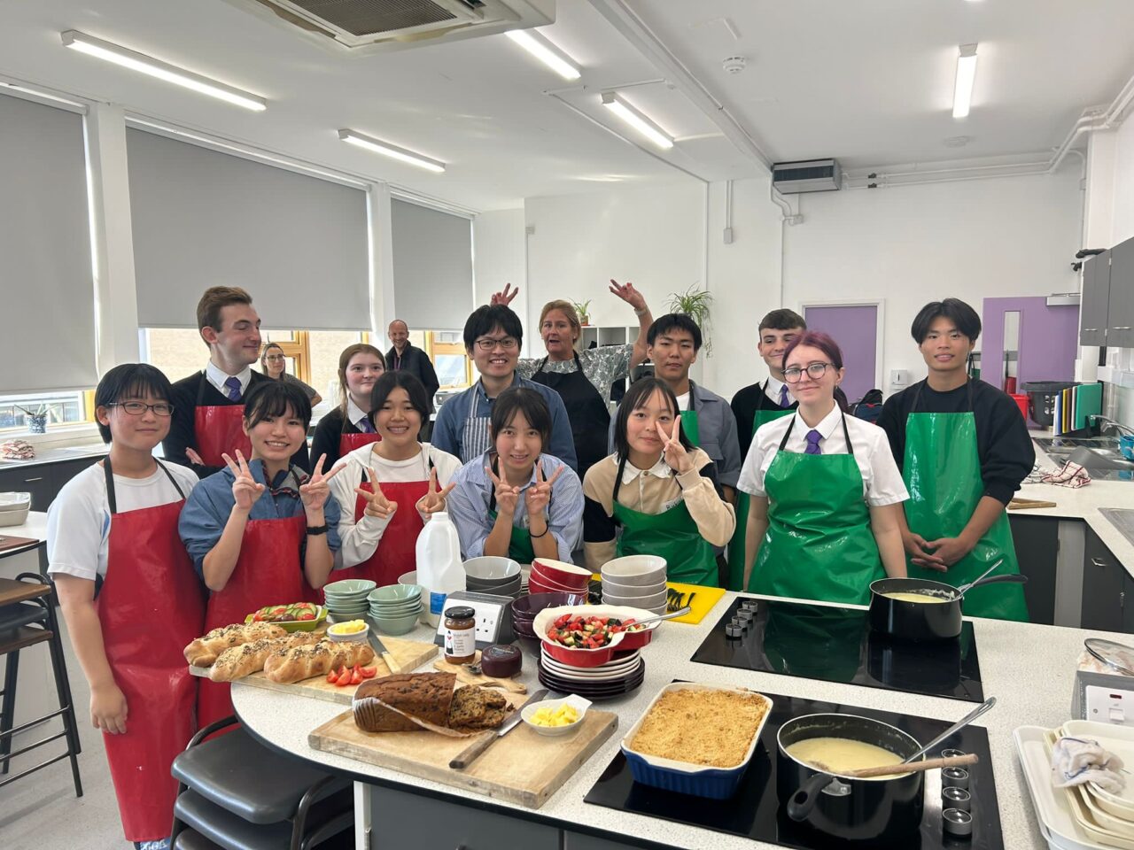 A group of girls and boys standing behind a cooking counter in a school classroom smiling at the camera. Some of the girls in the front of the group and a teacher at the back of the group, are doing the 'v' sign with their fingers. On the counter is a variety of locally baked Welsh food and bools and kitchen utensils. A tray of crumble and a saucepan of custard stands on the hob in the foreground.