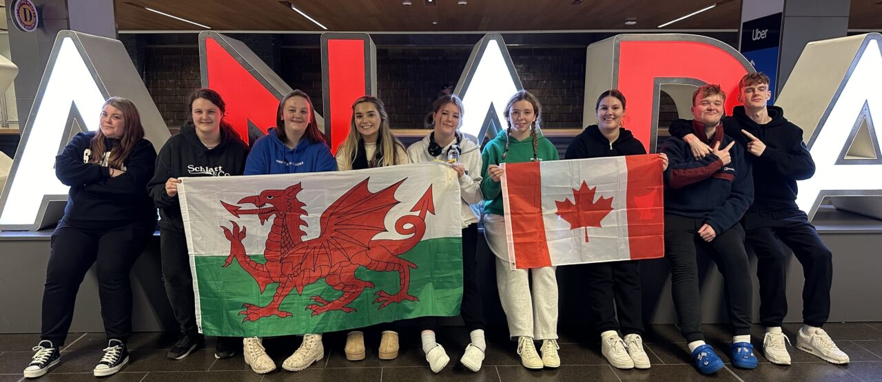 Myfyrwyr yn cyrraedd Canada, yn eistedd o flaen arwydd Canada yn dal baner Cymru a Chanada.