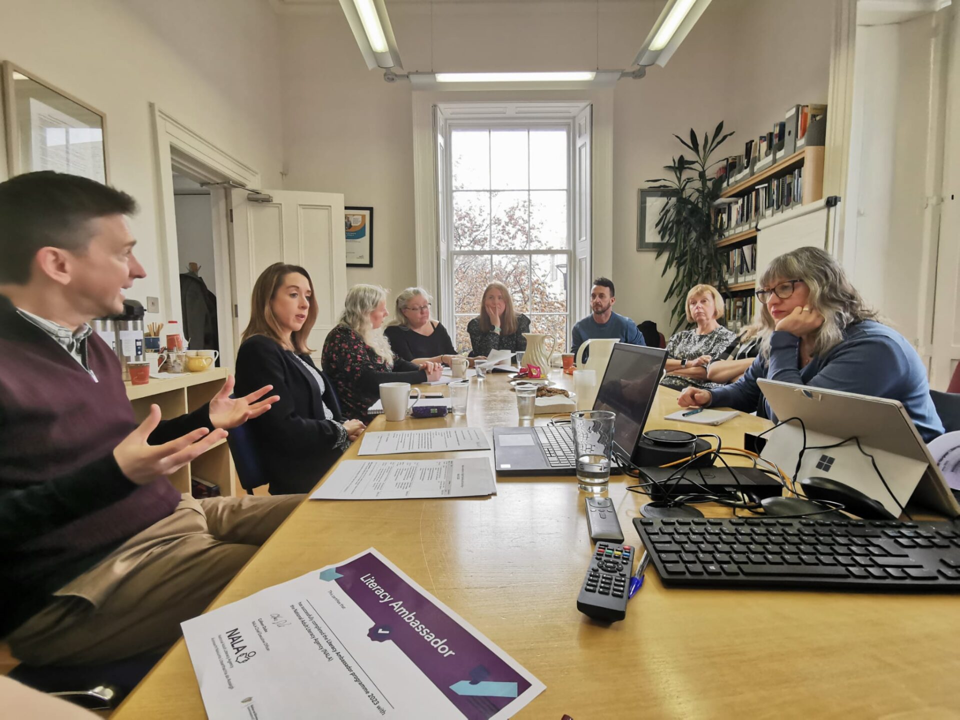 A group of people sat around a board table having a discussion. There are computers, papers and drinks on the table.