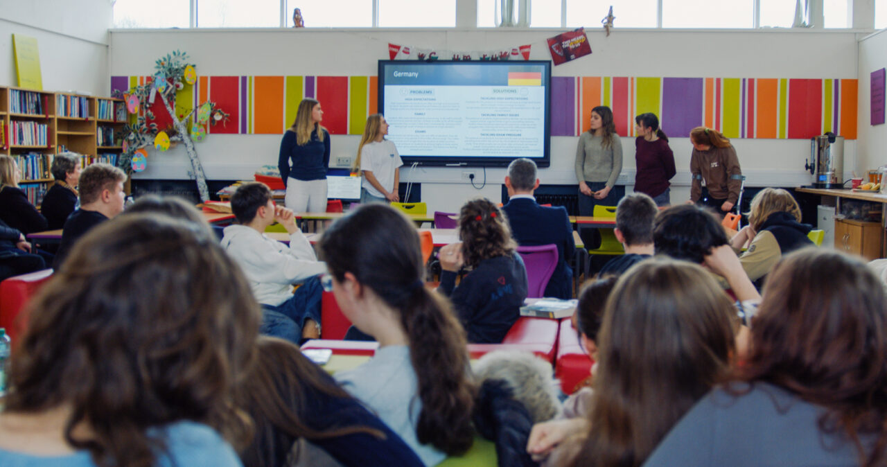 Five students stood at the front of a class presenting using a large TV screen. On the presentation you can see the word Germany and the German flag. In the foreground there is a class room full of pupils and staff sat facing the presentation.