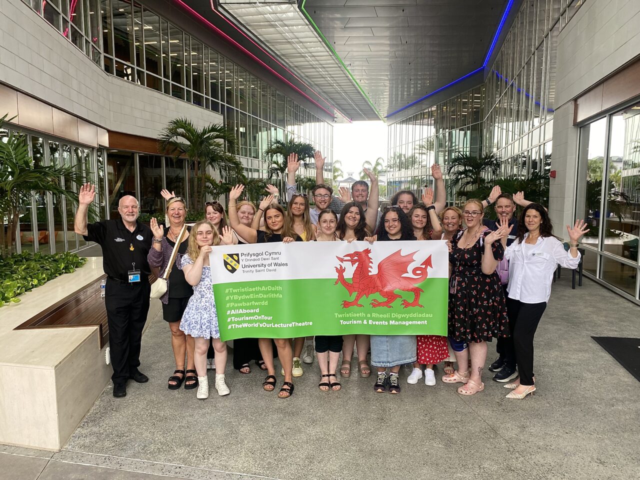 Student and staff from UWTSD standing in a large foyer space posing for a photo and holding a large banner with the Welsh flag and UWTSD on it.