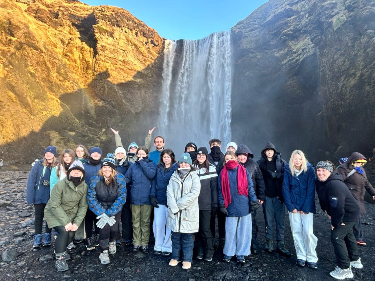 A group of young people and youth workers standing together at the base of a large waterfall in Iceland, surrounded by rocky cliffs and bright sunlight.