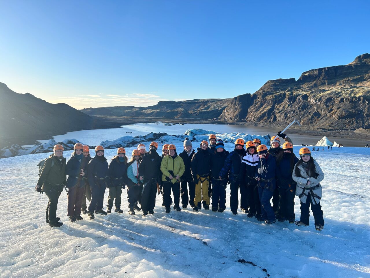 A group of participants wearing helmets and outdoor gear standing on a glacier in Iceland, with icy terrain and mountain scenery in the background under a clear blue sky.