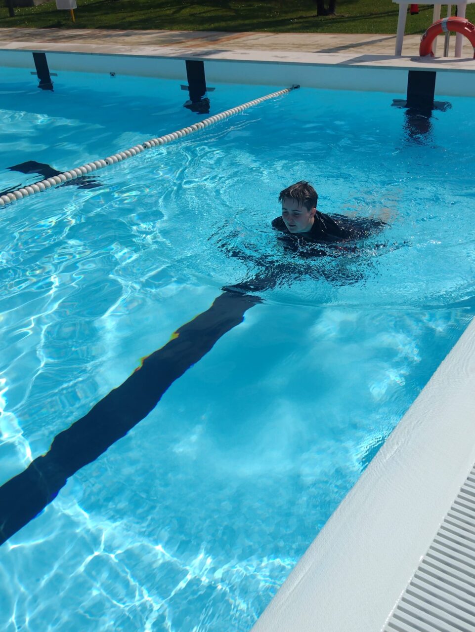 Paige, Gower College Student, in an outdoor swimming pool, partially submerged in clear blue water near a lane divider, with sunlight reflecting across the pool surface.