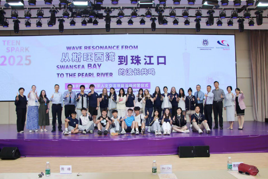 Group photo on a stage at the Teen Spark 2025 programme launch. Participants and staff stand and sit in front of a large screen displaying “Wave Resonance from Swansea Bay to the Pearl River” in English and Chinese.