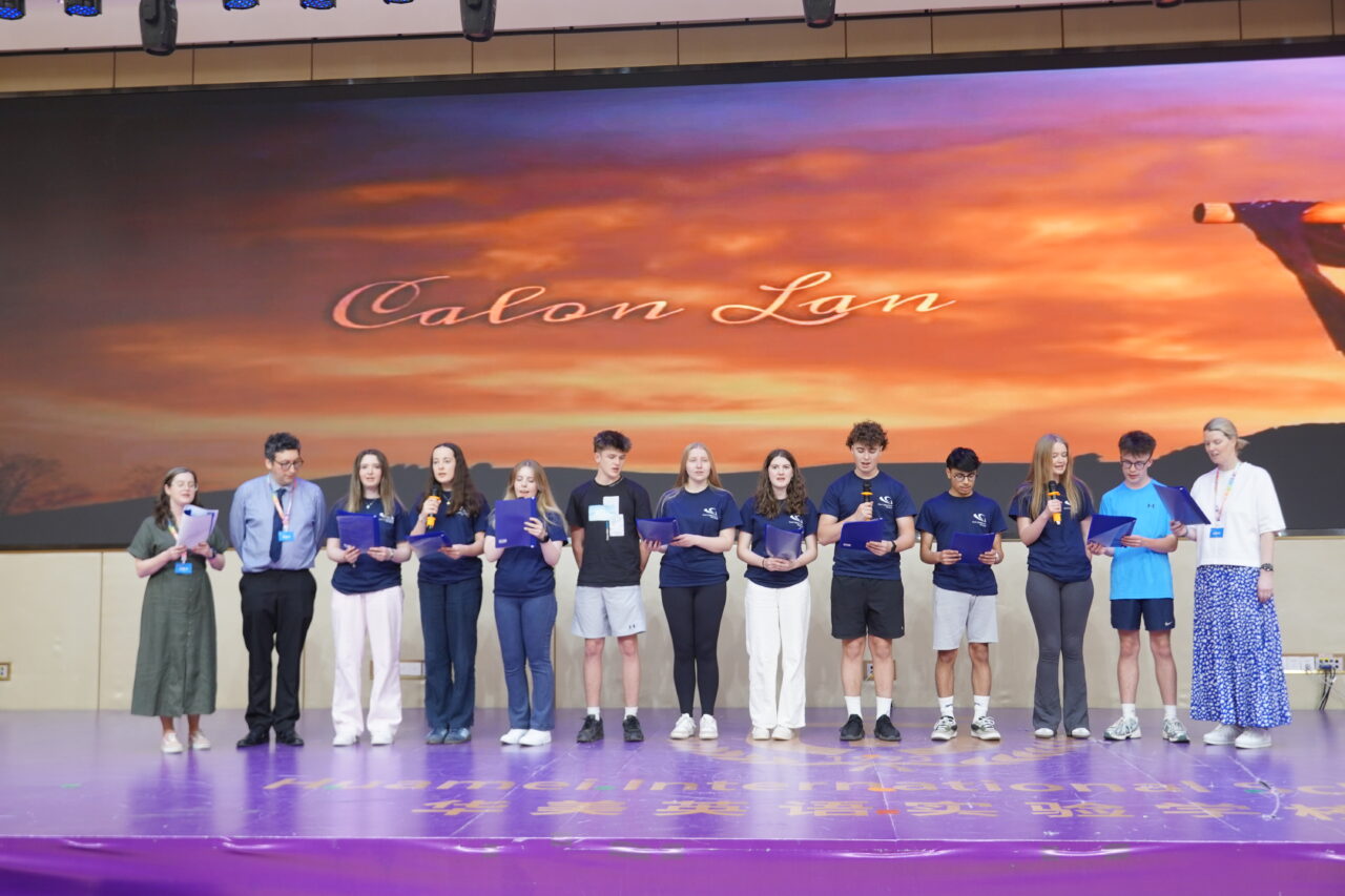 Participants and facilitators stand on stage reading from folders during a presentation at Teen Spark 2025. A large screen behind them displays the words “Calon Lan” against a sunset-themed background.