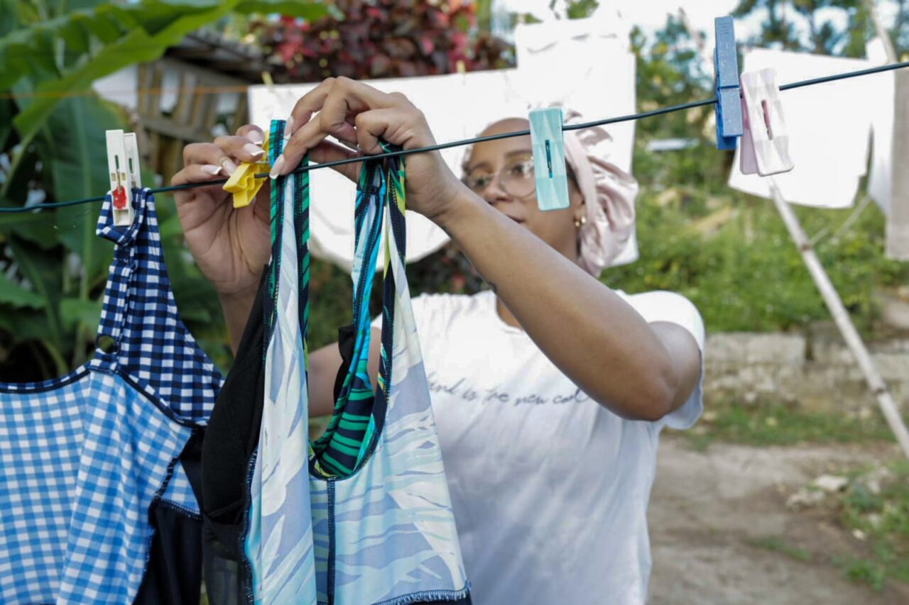 Person hanging patterned clothing on an outdoor washing line using colourful pegs, with greenery and laundry in the background at the Rastafari Indigenous Village
