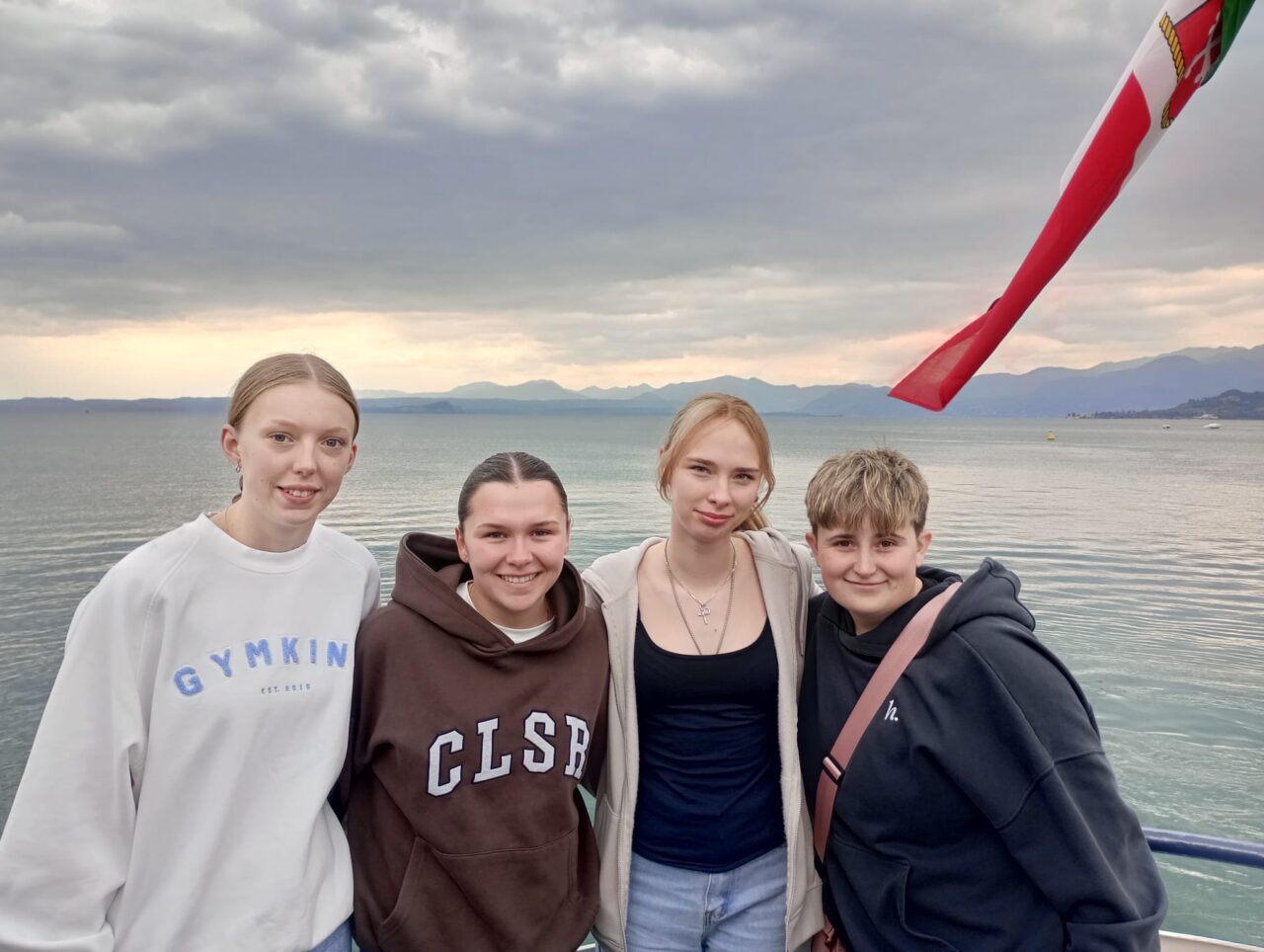 Paige, Gower College Student and three young people standing together on a waterfront promenade, with a calm lake, distant mountains, and cloudy skies in the background.
