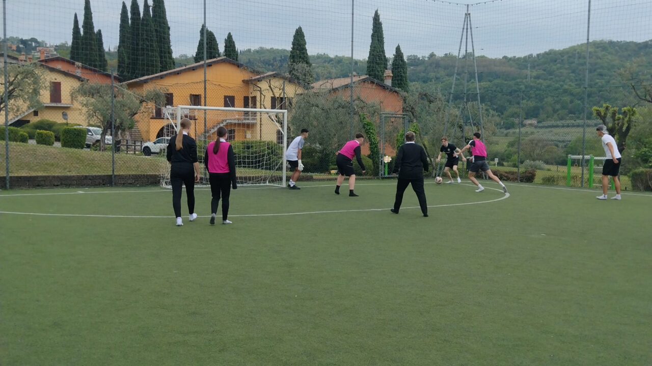 A group of young people playing football on an outdoor artificial turf pitch, with a goal, netted fencing, trees, and buildings in the background.