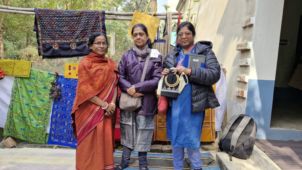 An outdoor craft display featuring colourful handmade textiles and embroidered bags. Three people stand in front of the display, with a variety of patterned fabric items hanging on racks behind them and displayed around the entrance of a building.