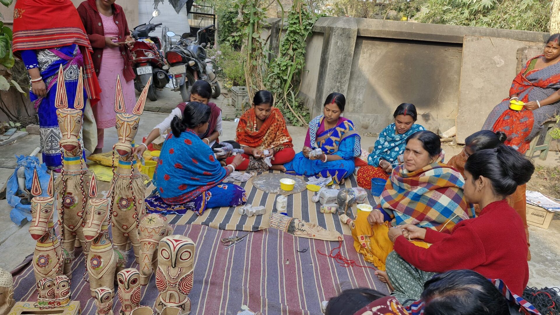 People gathered in an outdoor workspace making traditional Bengali decorative crafts. Painted figures with red, white, and natural-toned patterns stand at the front, while participants sit in a circle using craft materials and tools on mats.