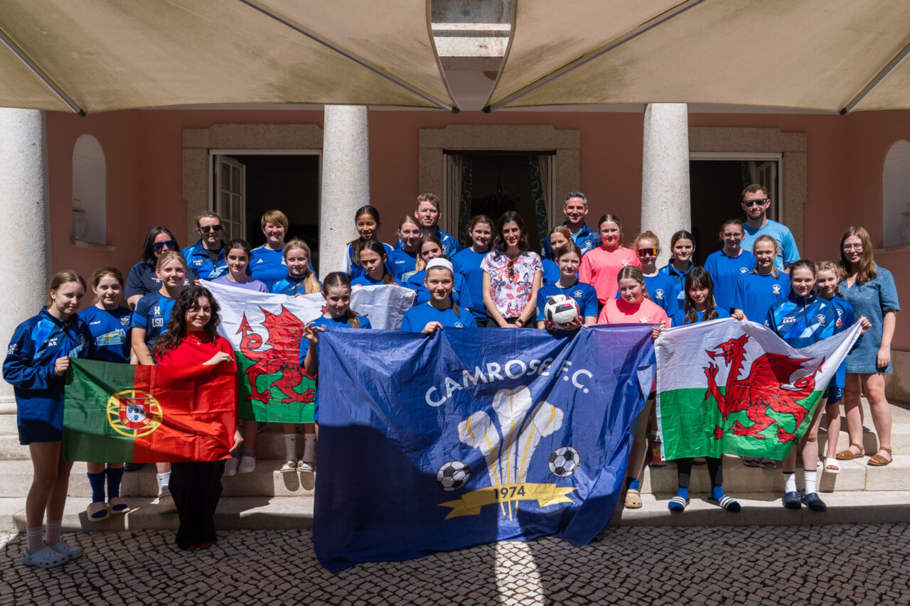 A group of young football players and coaches from Camrose AFC stand outside a building, holding Welsh, Portuguese and club flags during an international sports exchange visit.