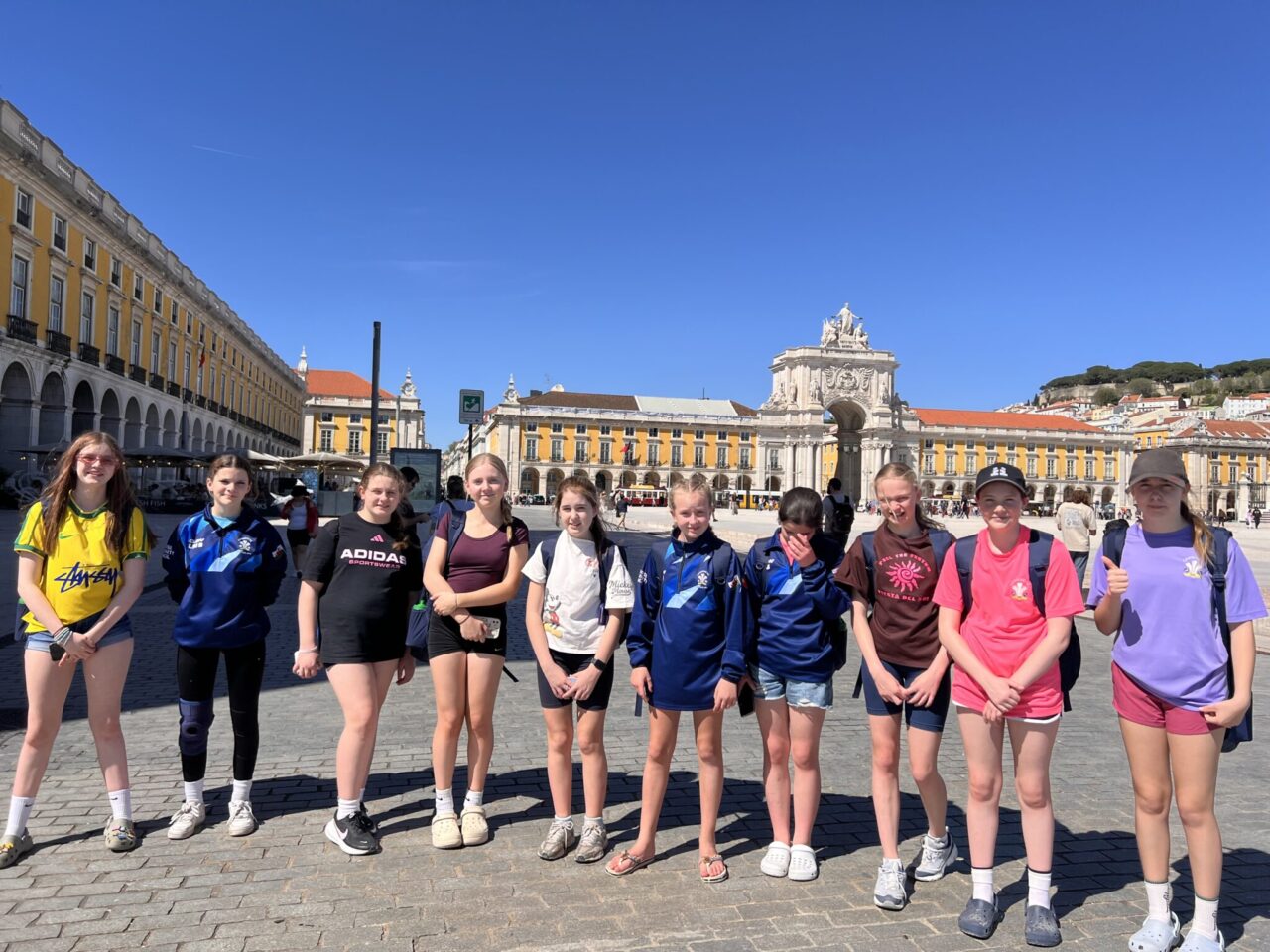 Young participants from Camrose AFC on a Taith-supported international exchange visit stand in Lisbon's Praça do Comércio, with the Arco da Rua Augusta archway and surrounding historic buildings behind them.