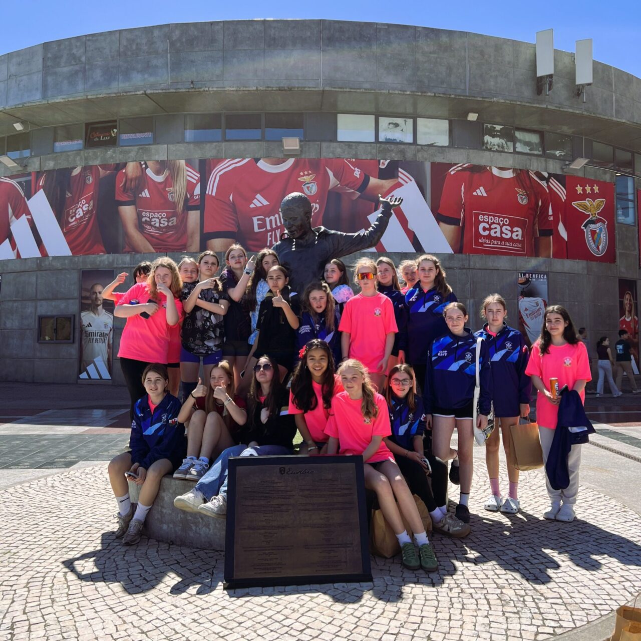 A group of young football players from Camrose AFC pose around a statue outside a football stadium, wearing team training kits during an international exchange visit.