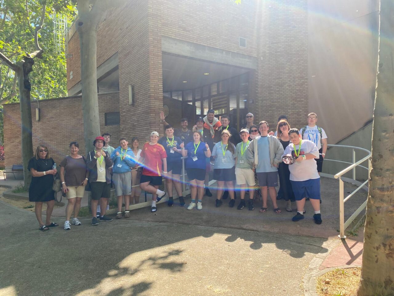 Group of young people and supporting adults standing outside a brick building surrounded by trees, posing together during a visit or group activity.