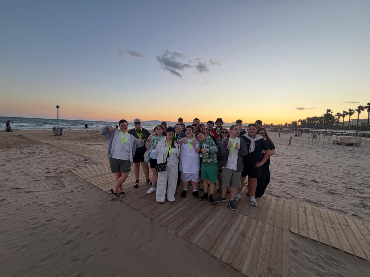Group of participants standing together on a wooden boardwalk on a sandy beach at sunset, with the sea on one side and palm trees in the distance. The group is wearing event lanyards and posing for a photo against a clear evening sky.