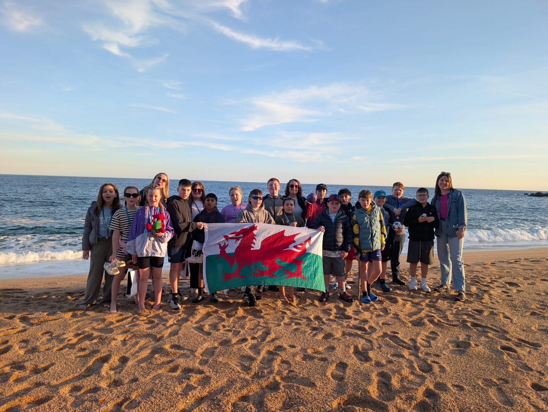 Group of Year 6 pupils from Ysgol Alexandra on a Barcelona beach holding a Welsh flag, taking part in a Taith-funded international mobility project.