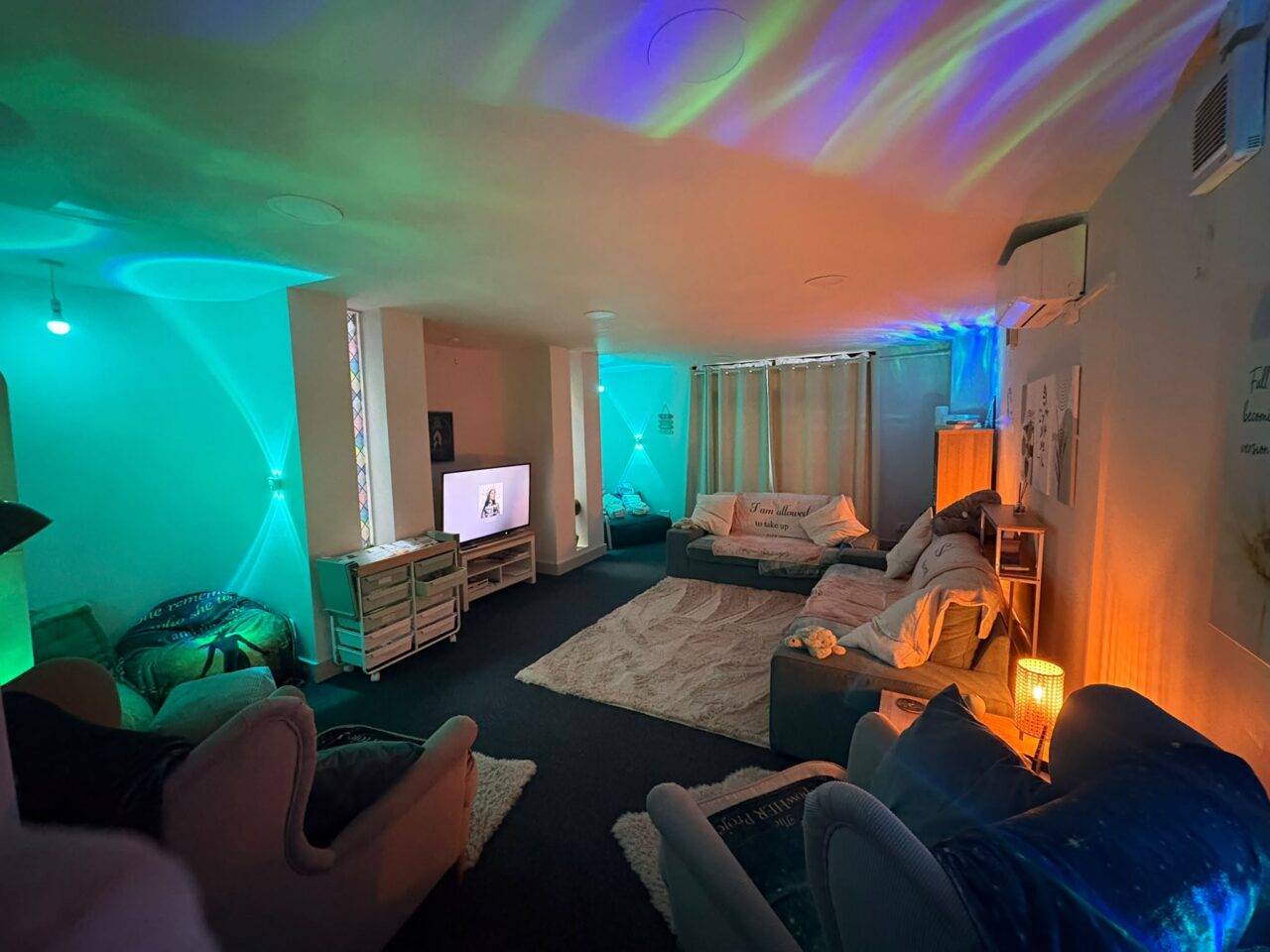 Calm wellbeing room with sofas, armchairs, rugs and soft ambient lighting. Colourful blue, green and orange light projections illuminate the walls and ceiling, creating a relaxing atmosphere.