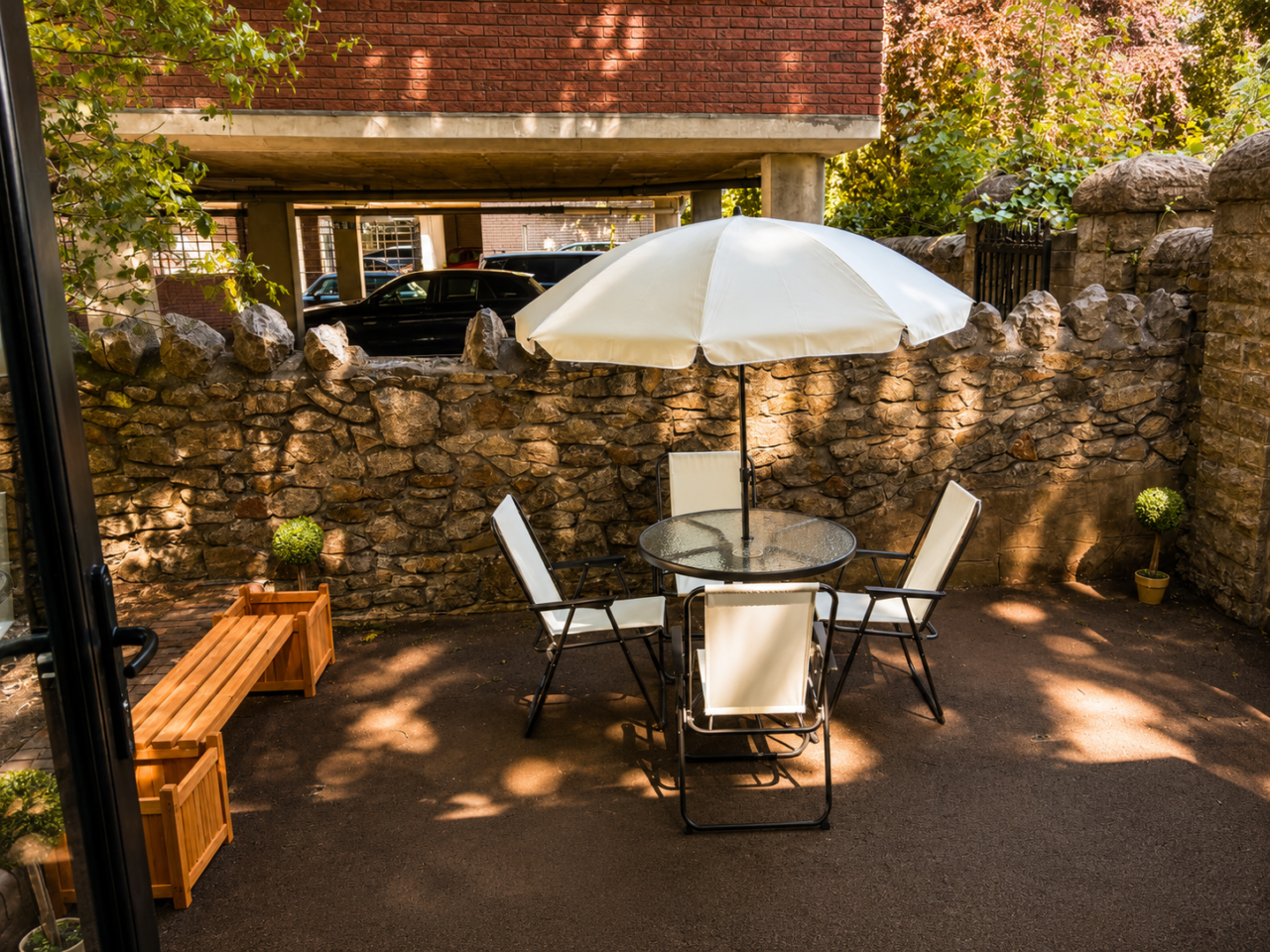 Private outdoor courtyard with a patio table, four chairs and a large white parasol. The enclosed space is bordered by stone walls and shaded by surrounding trees, creating a quiet area for relaxation and conversation.
