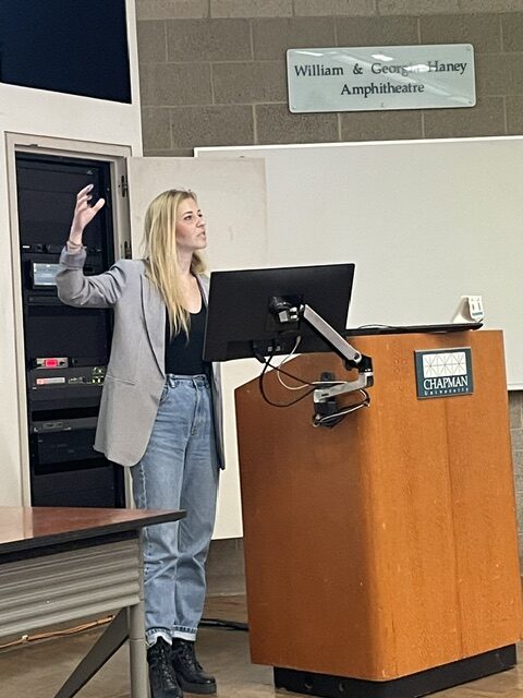 Marlen Komorowski presenting from a lectern in a university lecture theatre, gesturing while speaking to an audience.