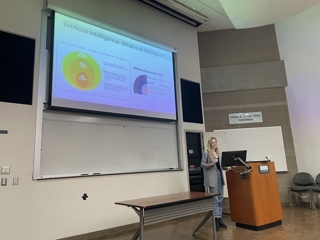 Marlen Komorowski delivering a presentation in a lecture theatre, standing beside a screen displaying slides about artificial intelligence.